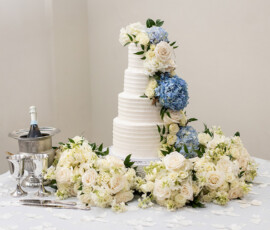 Elegant four-tier white wedding cake with white roses, blue hydrangeas, goblets, and champagne on a floral table.