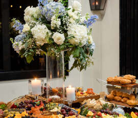 Elegant wedding buffet with cheeses, fruits, pastries, candles, and a white-blue floral arrangement in a glass vase.