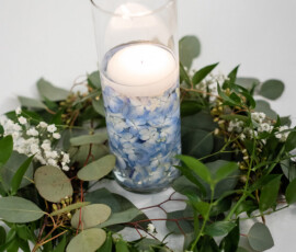 Elegant centerpiece with a glass vase, white floating candle, blue hydrangea petals, greenery, and small white flowers.