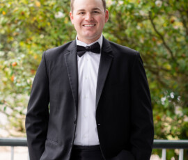 Smiling man in a black tuxedo with a bow tie stands outdoors, hands in pockets, green trees in background.
