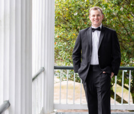 Man in black tuxedo smiles on a porch with white columns, green trees behind—classic New Orleans wedding charm.