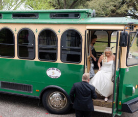 Bride in white dress exits green trolley with a suited man’s help; another woman and greenery show a classic New Orleans wedding.