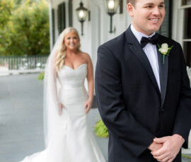 A groom in a black tuxedo smiles at his bride in a white gown and veil on a porch during their Louisiana wedding first look.