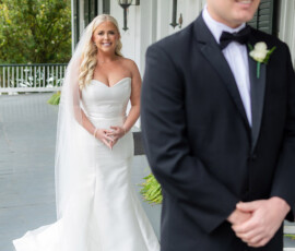 Bride in white gown smiles behind groom in black tuxedo on porch, joyful Louisiana wedding moment.