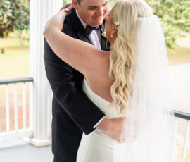 Bride in white dress and groom in black tux embrace on a porch at their New Orleans wedding, greenery in the background.