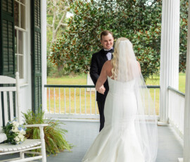 Bride and groom smile at each other on a porch at a Louisiana wedding, with bouquet on rocking chair and greenery behind.