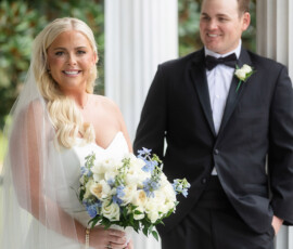 Bride in white with blue and white bouquet stands smiling beside groom in black tuxedo on a white-columned porch.