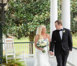 Bride in white dress and groom in black tuxedo smile on porch, holding hands at their New Orleans wedding.