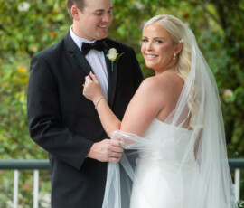Bride in white dress and veil smiles at camera, holding groom’s arm outdoors amid green foliage at their Louisiana wedding.
