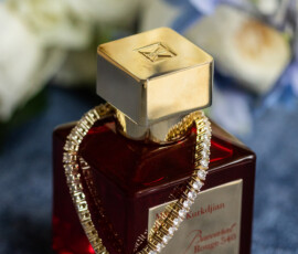 Red perfume bottle with gold accents and bracelet, blurred white and blue wedding flowers in the background.