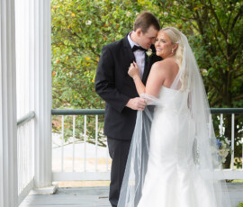 Bride in white gown and groom in black tuxedo embrace on porch with white columns, classic New Orleans wedding scene.