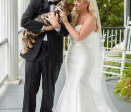 Bride and groom pet a small brown dog on a porch with white railings and greenery, Gulf South wedding charm.