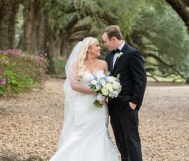 Bride in white dress and veil with bouquet stands beside groom in tux under arching trees on a leafy path, smiling at each other.