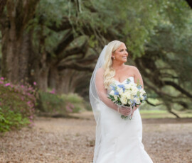 Bride in white dress with veil and blue-white bouquet stands on leaf-covered path under arching trees in New Orleans.