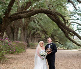Bride and groom in wedding attire stand on a leafy path, smiling with a bouquet and small dog under New Orleans trees.