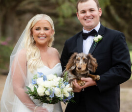 Bride and groom in wedding attire smile outdoors, holding a small dog and bouquet with white and blue flowers; trees behind them.
