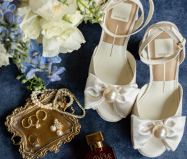 White sandals, gold jewelry, a red perfume bottle, and white-blue flowers on blue fabric—New Orleans wedding flat lay.