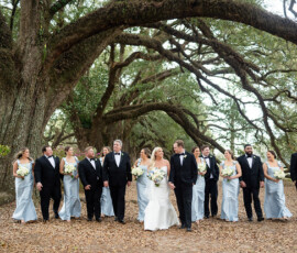 Bride, groom, and wedding party walk under arching Louisiana trees; bridesmaids in blue, groomsmen in black tuxedos, all smiling.