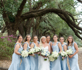Bridal party in pale blue and white dresses smiles outdoors under arching trees at a Louisiana wedding.