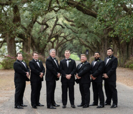 Seven men in black tuxedos pose on a tree-lined path at a Louisiana wedding, with oak trees and lush greenery in the background.