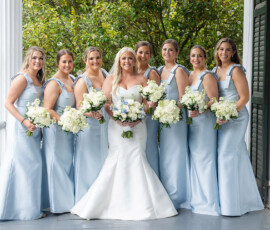 Bride with seven bridesmaids in pale blue dresses smile on a porch with bouquets, ready for their Gulf South wedding.