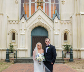 Bride in white gown and groom in tuxedo smile outside ornate Louisiana church after their wedding.
