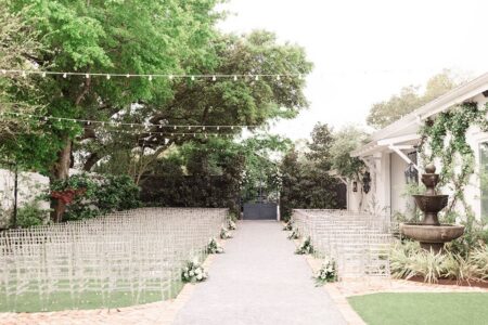 Outdoor wedding aisle with clear chairs, white flowers, green trees, string lights, and a fountain by a white building in New Orleans.