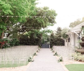 Outdoor wedding aisle with clear chairs, white flowers, green trees, string lights, and a fountain by a white building in New Orleans.