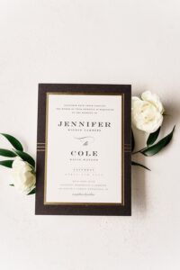 Elegant wedding invitation with black border, gold accents, white roses, green leaves; announces Jennifer and Cole’s marriage.