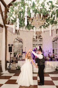 Bride and groom dance under a floral chandelier at their New Orleans wedding, with string lights and live band in the background.