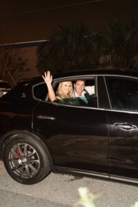 A smiling woman waves from a black car at night after a New Orleans wedding, with a man beside her and trees in the background.
