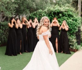 Bride in a white gown smiles at the camera as bridesmaids in black dresses stand behind her, eyes covered in anticipation.