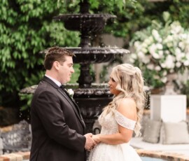 Bride and groom hold hands by a white-flowered fountain, lush greenery behind, at an elegant New Orleans wedding.