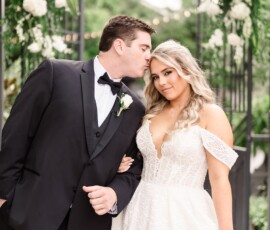 Groom in black tux kisses bride’s forehead. Bride wears off-shoulder lace gown. Smiling, they stand by floral wedding decor.