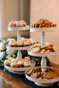 Three white tiered trays hold cookies and cupcakes on a wooden table with greenery, ideal for a Gulf South wedding reception.