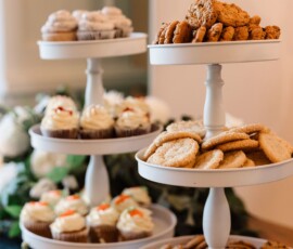 Three white tiered trays hold cookies and cupcakes on a wooden table with greenery, ideal for a Gulf South wedding reception.