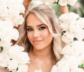 Smiling blonde woman with soft makeup framed by white rose bouquets, perfect Louisiana wedding inspiration.