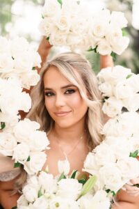 Smiling blonde woman with soft makeup framed by white rose bouquets, perfect Louisiana wedding inspiration.