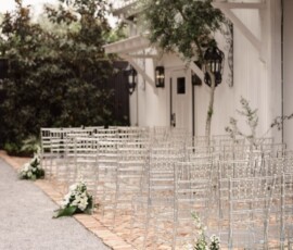 Clear chairs line a brick aisle with white florals by a white building, creating an elegant New Orleans wedding setting.