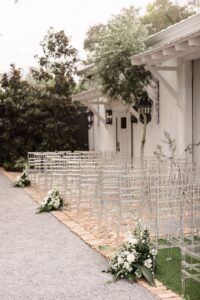 Clear chairs line a brick aisle with white florals by a white building, creating an elegant New Orleans wedding setting.