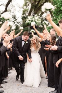 Bride and groom walk hand-in-hand, smiling, as guests in black cheer with white bouquets at a Louisiana wedding outdoors.
