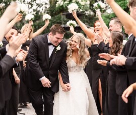 Bride and groom walk hand-in-hand, smiling, as guests in black cheer with white bouquets at a Louisiana wedding outdoors.