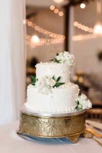 Elegant white two-tier wedding cake with roses, greenery, and pearls on a gold stand, set amid string lights.