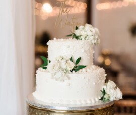 Elegant white two-tier wedding cake with roses, greenery, and pearls on a gold stand, set amid string lights.