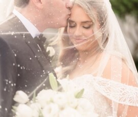 Groom kisses bride’s forehead under a sheer veil, both with closed eyes and white bouquet at their New Orleans wedding.
