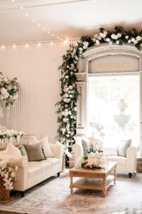 White room with sofas, coffee table, lush white flowers, arched window with SUPPOSEY sign, and glowing string lights.
