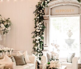 White room with sofas, coffee table, lush white flowers, arched window with SUPPOSEY sign, and glowing string lights.