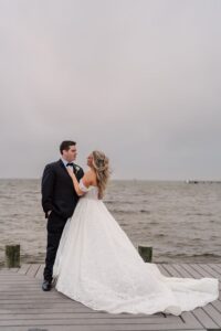 Bride in white gown and groom in black tuxedo share a romantic moment on a Louisiana dock under cloudy skies.