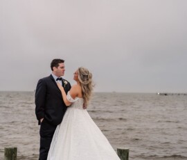 Bride in white gown and groom in black tuxedo share a romantic moment on a Louisiana dock under cloudy skies.