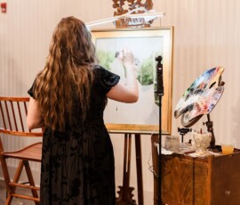 A woman in a black dress paints at an easel, inspired by a Gulf South wedding, with art supplies on a nearby table.